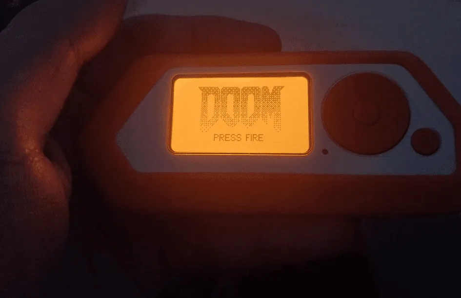 Photo of a hand holding a Flipper Zero showing the DOOM game with a Press Fire prompt on its screen.